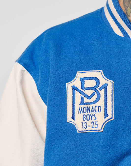 Close-up of the LIVE FAST DIE YOUNG x Schott NYC Varsity Jacket in blue and white, featuring cowhide leather sleeves and a chenille Monaco Boys 13-25 patch with a stylized MB logo on the chest.