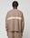 A person with short, twisted hair is seen from behind wearing the LIVE FAST DIE YOUNG Live Fast Boucle Sweater in light brown with "LIVE FAST" in white, paired with matching knit pants. The background is plain gray.