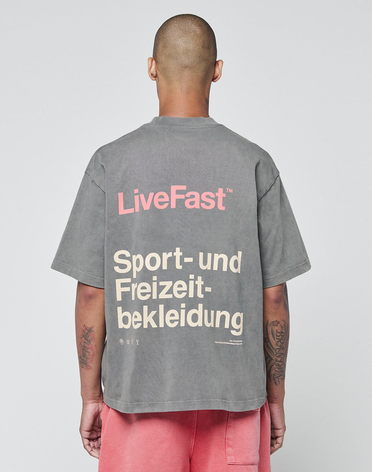 Seen from behind, a person with a shaved head wears the LIVE FAST DIE YOUNG Basic Trademark Tee in gray with pink LiveFast and beige Sport- und Freizeitbekleidung text. Oversize fit and pink shorts create a unique look against a plain background. | model