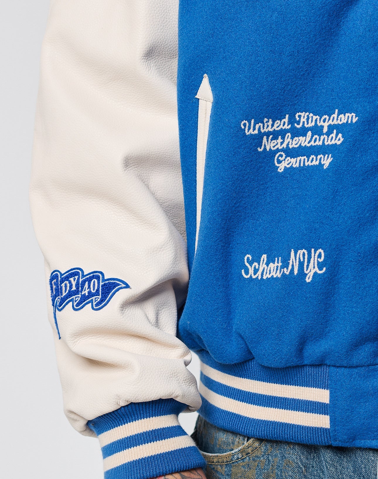 A close-up of the LFDY x Schott NYC Varsity Jacket by LIVE FAST DIE YOUNG, showing blue and white sleeves with chenille patches, a NY 40 patch, and embroidered United Kingdom, Netherlands, Germany, Schott NYC text.