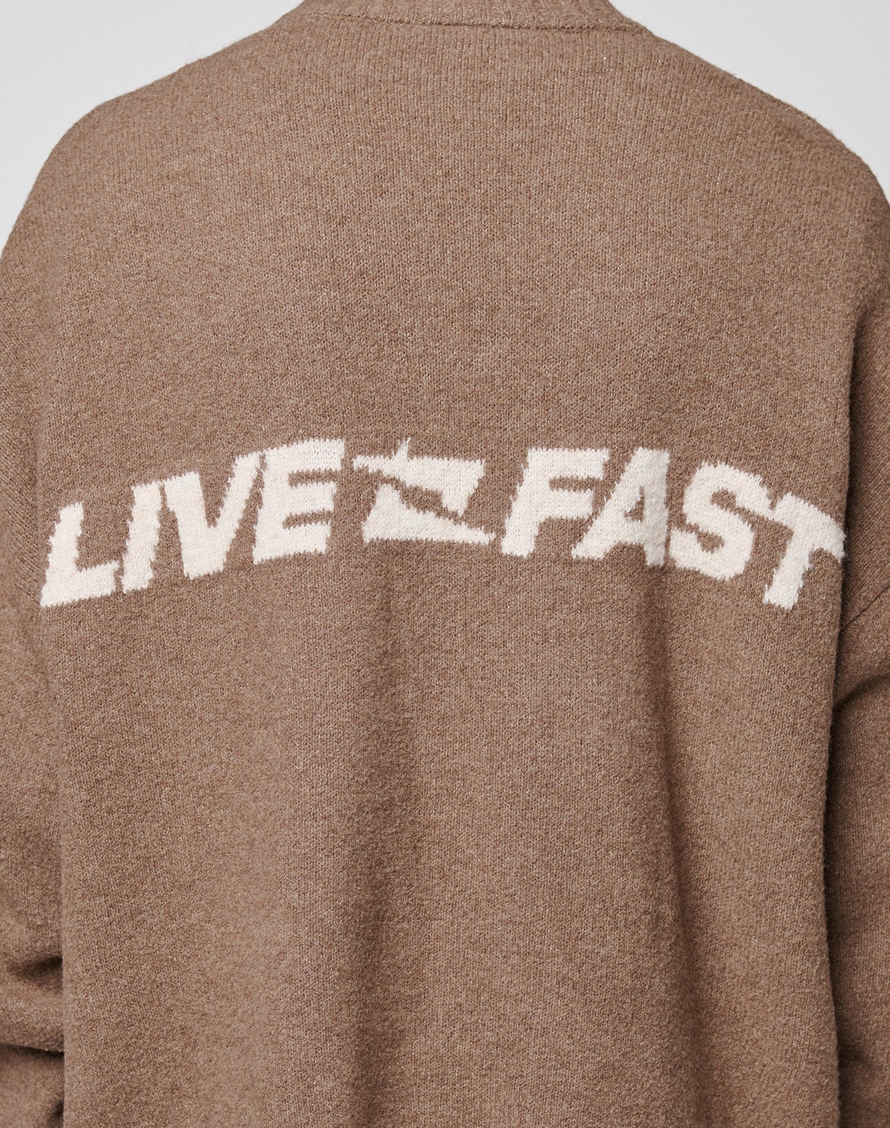 A close-up of the Live Fast Boucle Sweater by LIVE FAST DIE YOUNG shows its brown oversized fit, with "LIVE FAST" in large white block letters—A replaced by a lightning bolt—across the back for a bold effect.