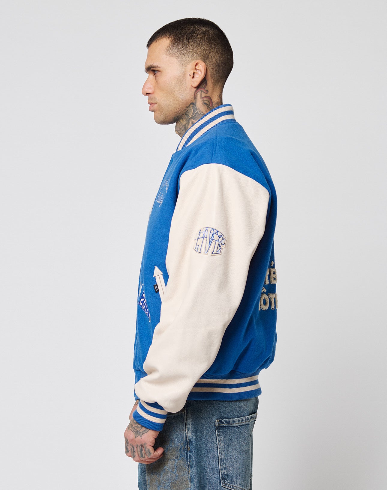 A tattooed man in profile wears the LIVE FAST DIE YOUNG LFDY x Schott NYC Varsity Jacket, featuring blue and white colors with cowhide leather sleeves and jeans, against a plain light gray background.
