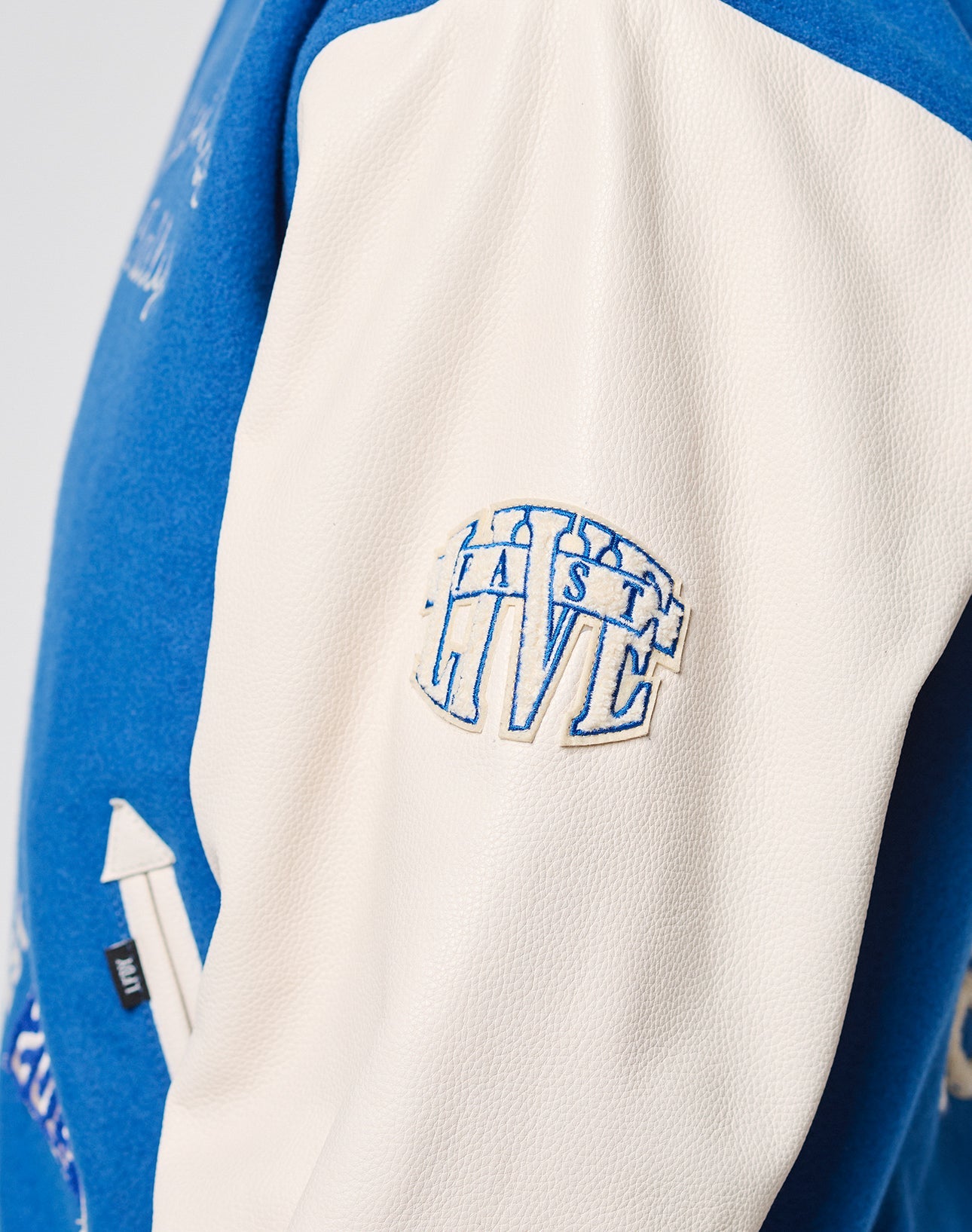 A close-up of the white cowhide leather sleeve on the LFDY x Schott NYC Varsity Jacket by LIVE FAST DIE YOUNG, with blue LAST LIVE embroidery on the upper arm.
