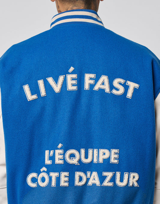 A person is seen from the back wearing the LFDY x Schott NYC Varsity Jacket by LIVE FAST DIE YOUNG, featuring blue and white colors, cowhide leather sleeves, and bold white text: LIVE FAST and LÉQUIPE CÔTE DAZUR.