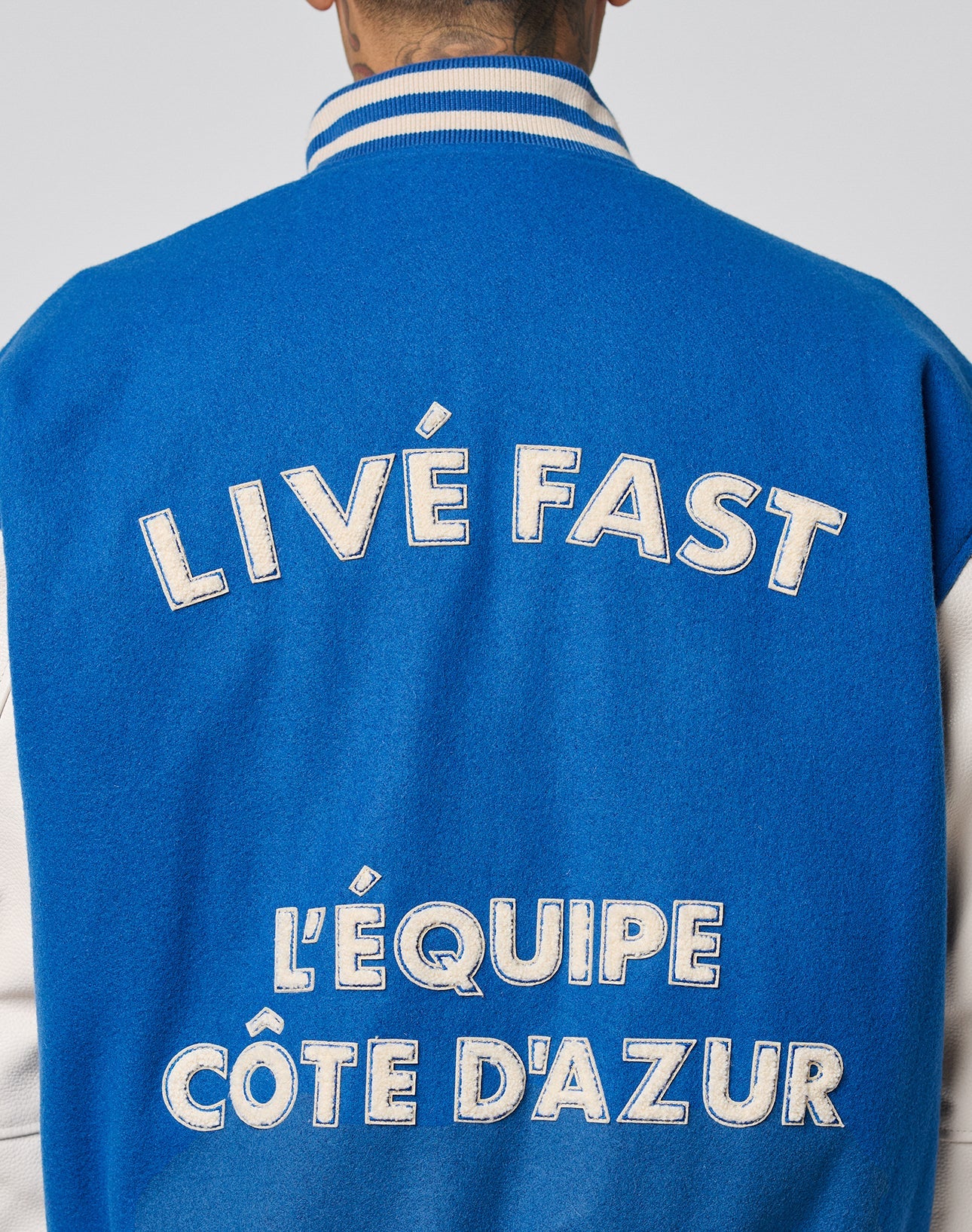A person is seen from the back wearing the LFDY x Schott NYC Varsity Jacket by LIVE FAST DIE YOUNG, featuring blue and white colors, cowhide leather sleeves, and bold white text: LIVE FAST and LÉQUIPE CÔTE DAZUR.