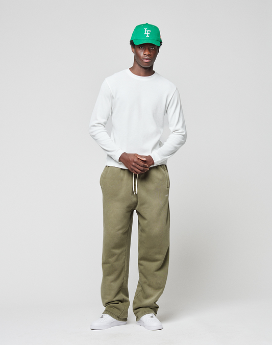A man stands against a plain background wearing a white long-sleeve shirt, LIVE FAST DIE YOUNG Basic Wide Jogger sweatpants in olive green, white sneakers, and a green baseball cap.