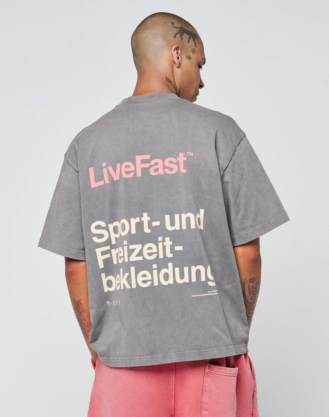 A tattooed person with a shaved head faces away, wearing a gray LIVE FAST DIE YOUNG Basic Trademark Tee featuring front and back print, paired with pink shorts for an oversized fit.