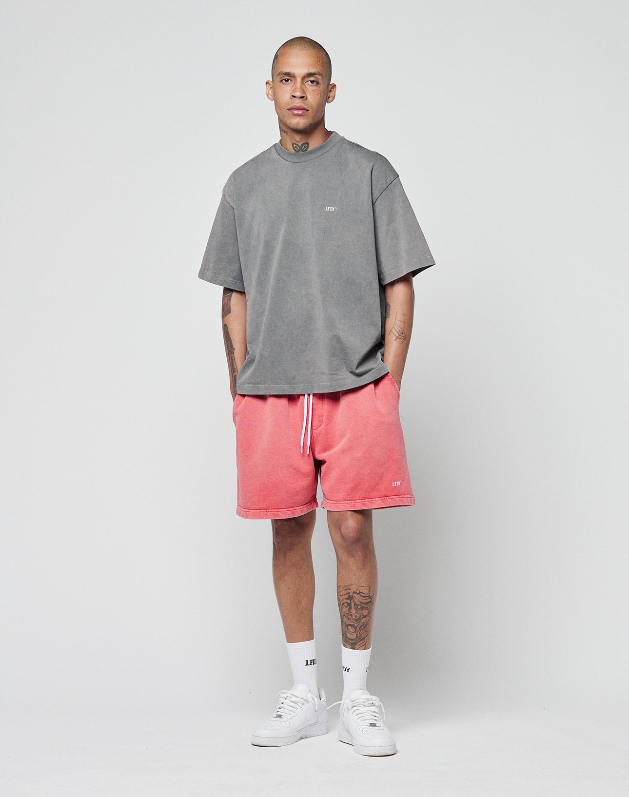 A man with tattoos stands facing forward against a plain light gray background, wearing a LIVE FAST DIE YOUNG Basic Trademark Tee in gray, paired with pink drawstring shorts, white socks, and sneakers.