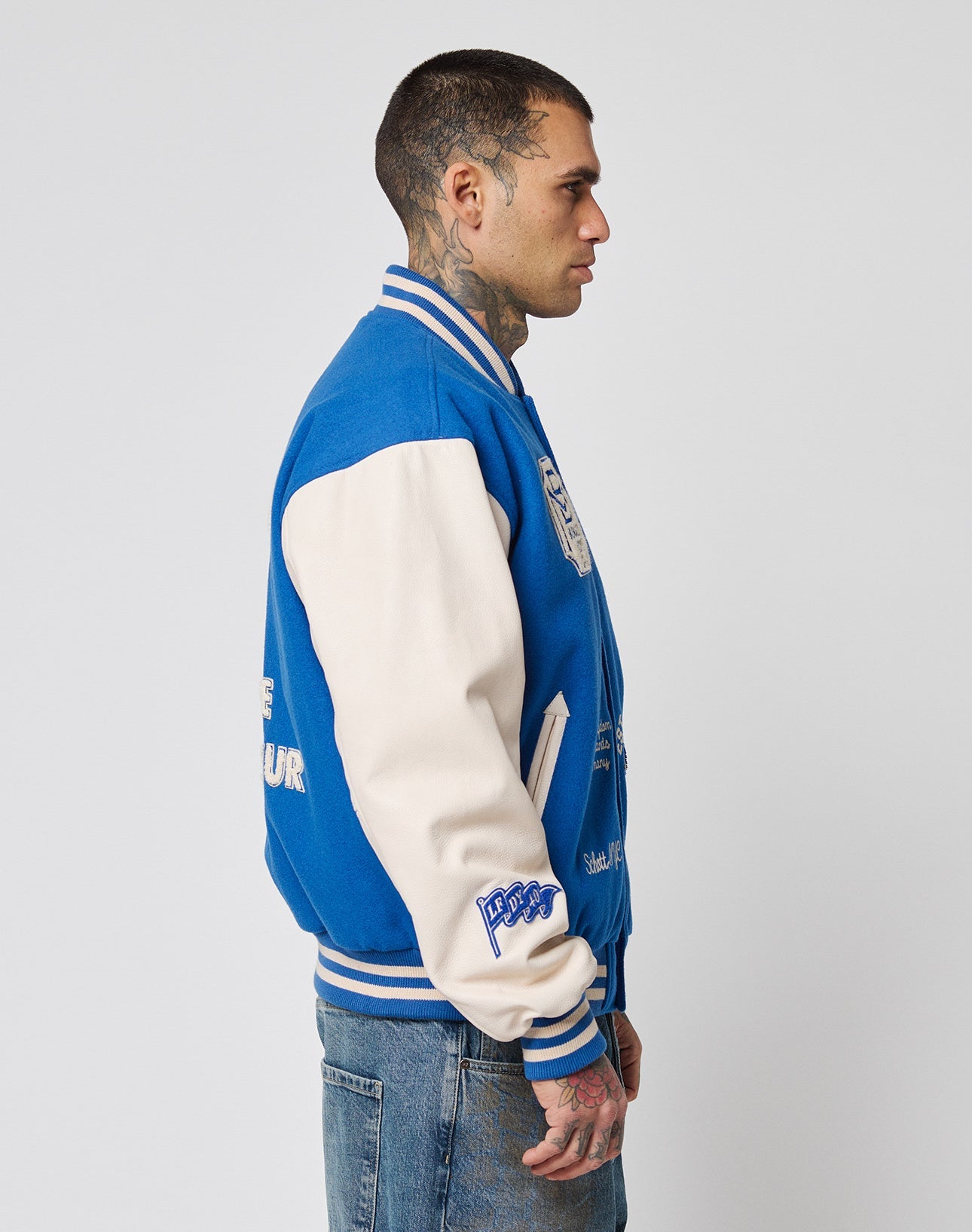 A tattooed man in jeans and short hair stands in profile against a plain background, wearing the LIVE FAST DIE YOUNG LFDY x Schott NYC Varsity Jacket with cowhide leather sleeves and chenille patches.