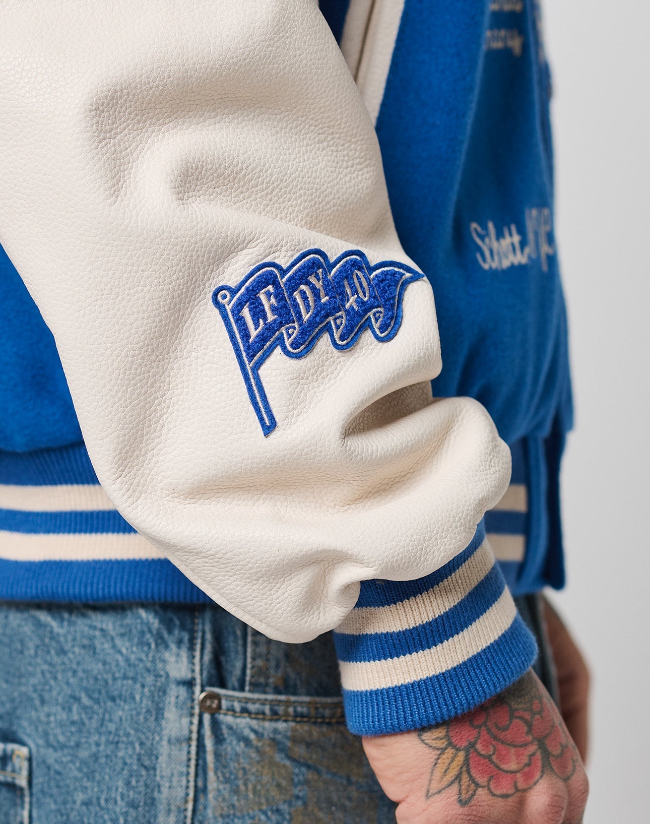 Close-up of a person’s arm in the LFDY x Schott NYC Varsity Jacket by LIVE FAST DIE YOUNG, featuring blue and white colors, chenille patches, cowhide leather sleeves, a signature flag patch, colorful flower tattoo, and blue jeans.