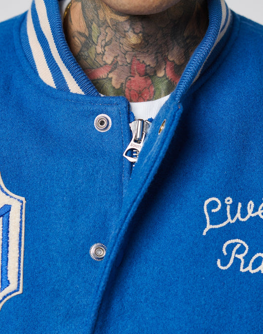 Close-up of a person in a LIVE FAST DIE YOUNG LFDY x Schott NYC Varsity Jacket with chenille patches, cowhide leather sleeves, white trim, and embroidered text. Colorful floral neck tattoos are visible above a white shirt.