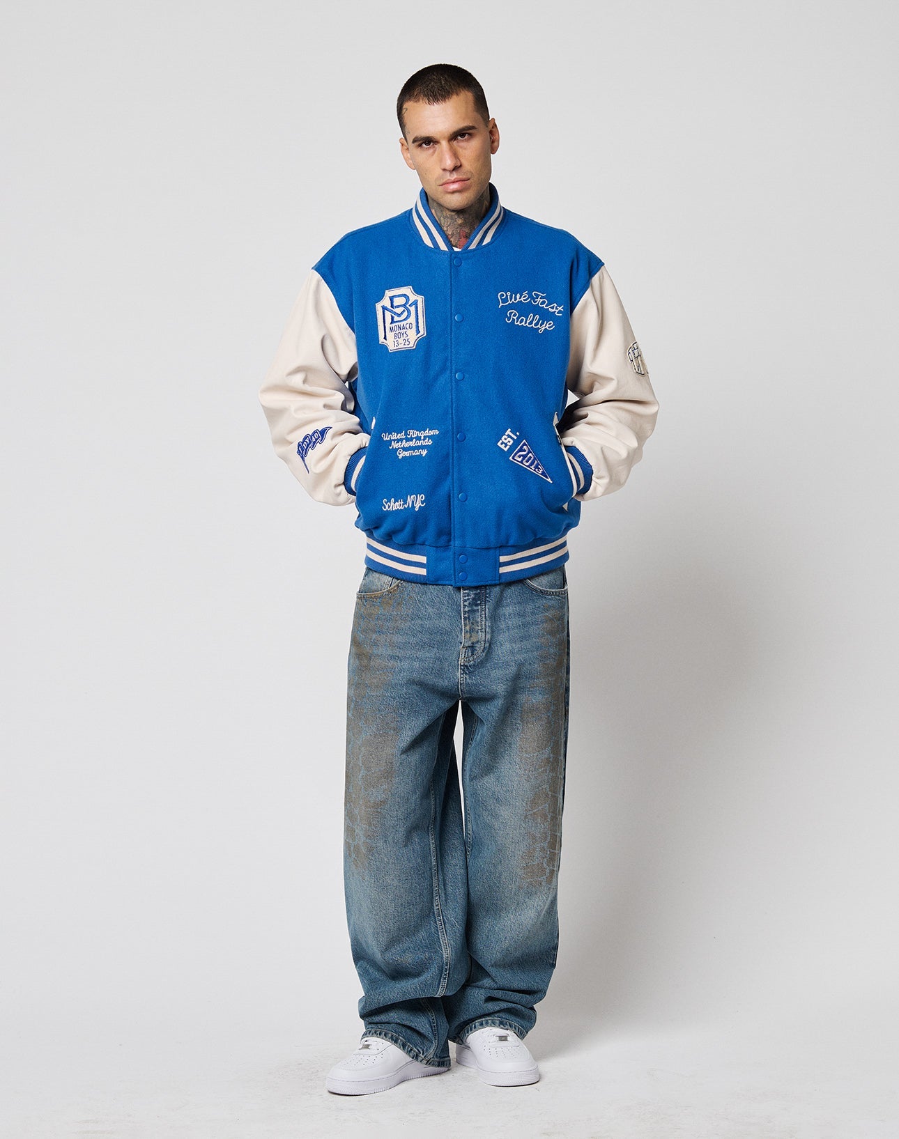 A man stands in a studio facing forward, wearing the LIVE FAST DIE YOUNG LFDY x Schott NYC Varsity Jacket with cowhide leather sleeves, loose jeans, and white sneakers against a plain white background.