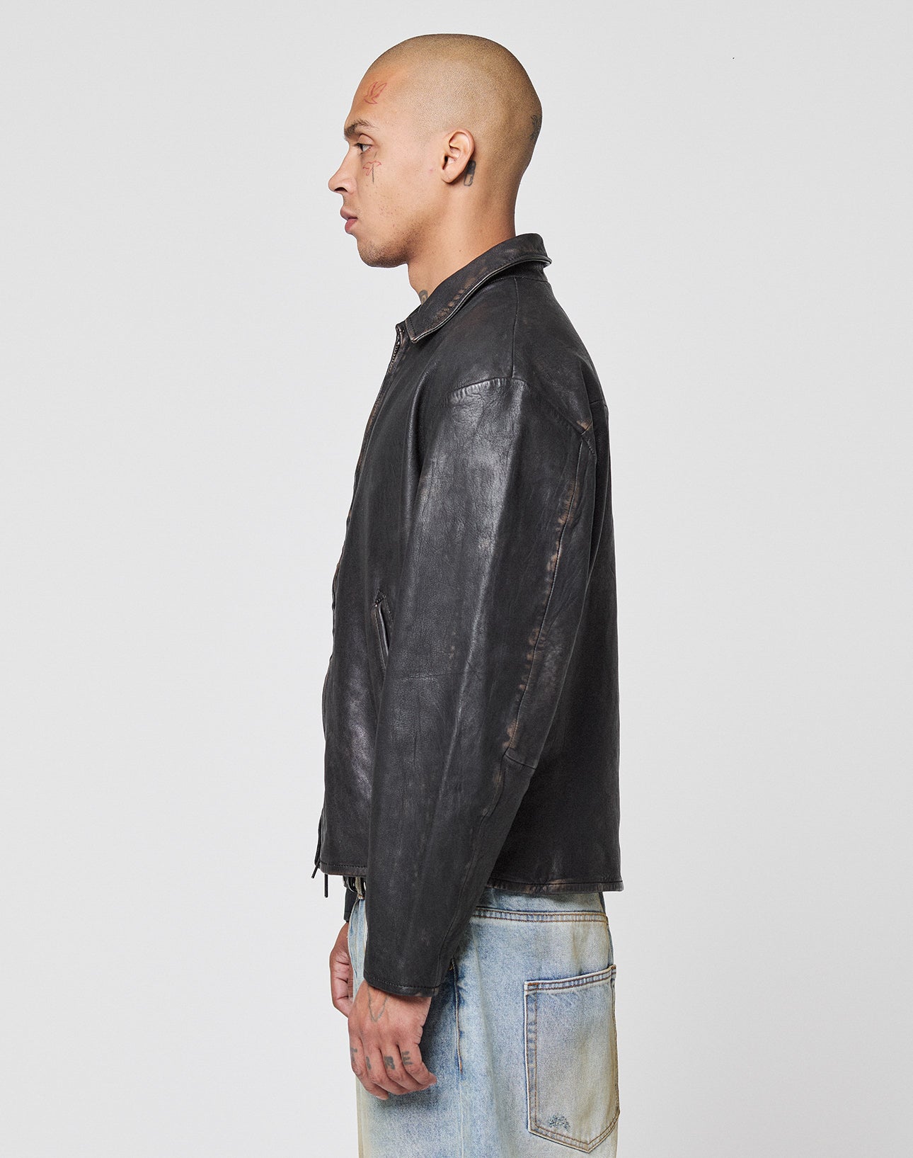 A person with a shaved head, wearing a black LIVE FAST DIE YOUNG LFDY Leather Jacket and light blue jeans, stands in profile against a plain white background.