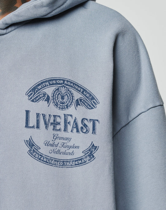 A close-up of the light blue LFDY 55 Hooded Zip by LIVE FAST DIE YOUNG in Oversize Fit, featuring a navy LIVE FAST graphic with Germany, United Kingdom, Netherlands, text banners, winged emblem, and stylish vintage wash.
