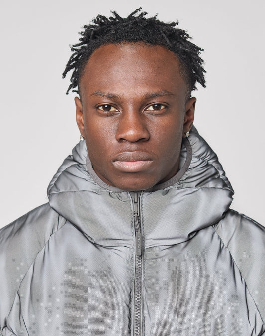 A person with short, twisted hair wears a Live Fast Puffer Jacket by LIVE FAST DIE YOUNG, hood up, and looks directly at the camera with a neutral expression against a plain light background.