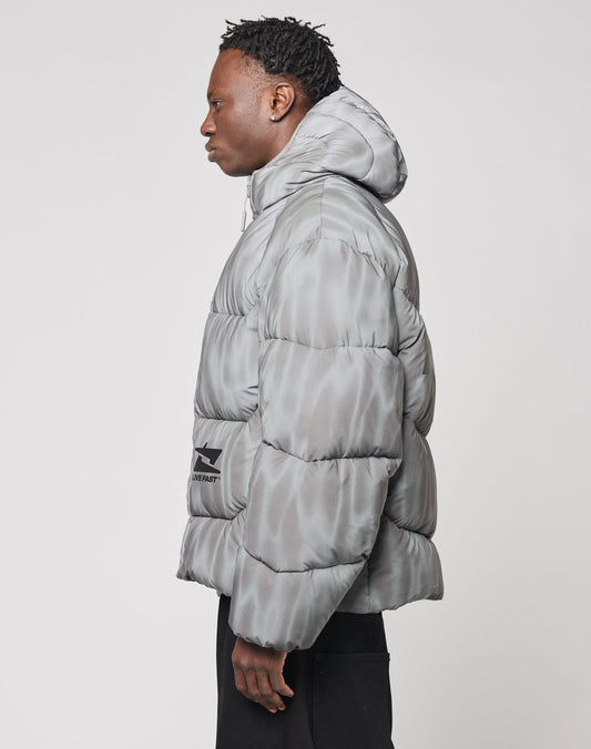 A person in profile models the Live Fast Puffer Jacket by LIVE FAST DIE YOUNG—an oversize, light gray, water-repellent puffer with subtle pattern and branded piping on the hood—paired with black pants against a plain white background.