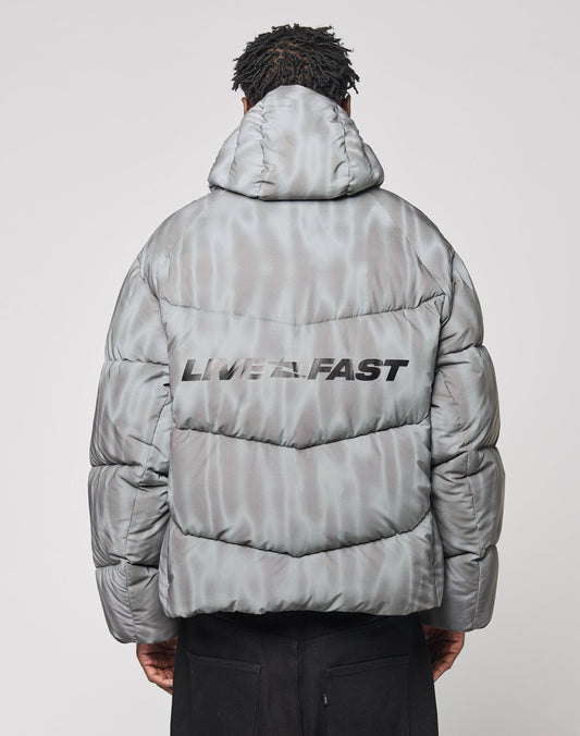 A person with their back to the camera wears the gray LIVE FAST DIE YOUNG Live Fast Puffer Jacket, featuring a hood with branded piping and bold black LIVE FAST lettering on the back.