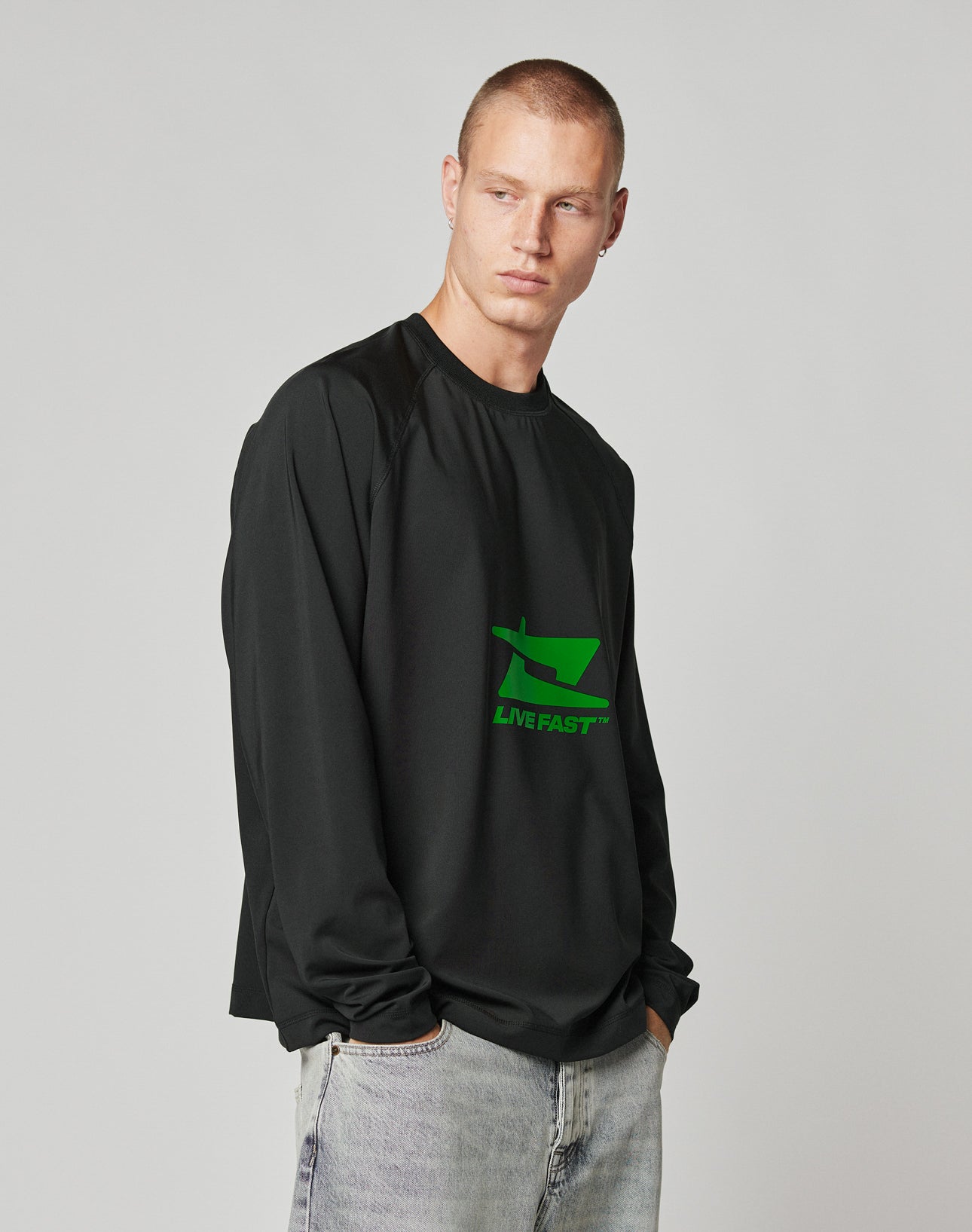 A man with short hair wears the LFDY Sports Longsleeve by LIVE FAST DIE YOUNG, featuring Raglan sleeves and a bright green LIVE FAST™ graphic, paired with light-wash jeans against a plain light gray background.