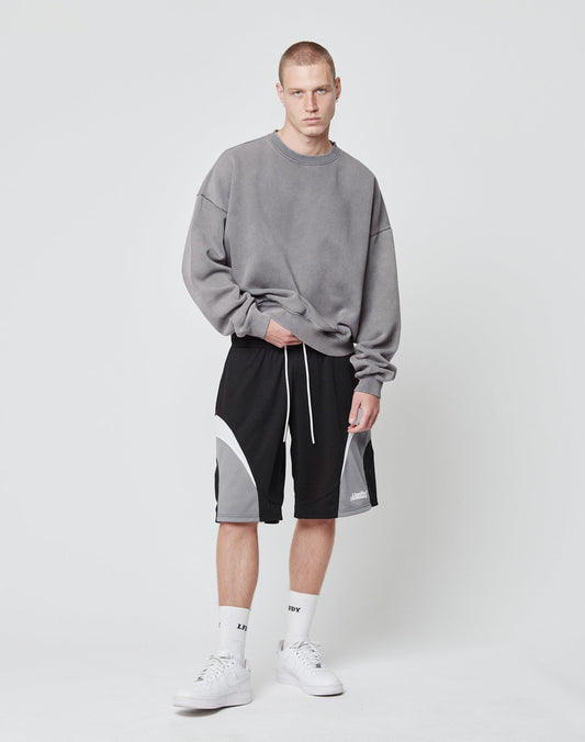 A young man with short hair and a neutral expression stands against a white background, wearing LIVE FAST DIE YOUNG Basic Trackshorts with an elastic waistband, a gray oversized sweatshirt, white socks, and white sneakers.