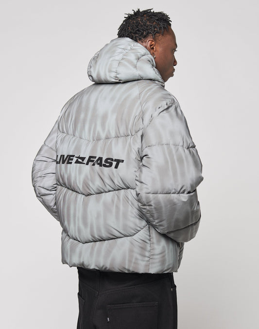 A person is seen from the back wearing the Live Fast Puffer Jacket by LIVE FAST DIE YOUNG in light gray with a water-repellent finish, branded hood piping, and LIVE FAST boldly printed across the upper back. The jacket has an oversized fit.
