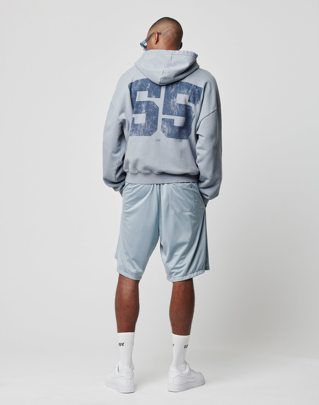 A person faces away, wearing a light blue oversize hoodie with a large 55 on the back, LIVE FAST DIE YOUNG LFDY Loyal Shorts with elastic waistband, white Nike socks, and white sneakers against a plain white background.