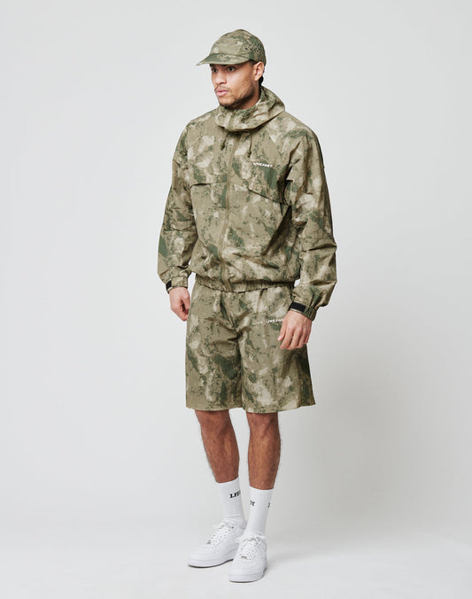 A man stands against a plain background wearing LIVE FAST DIE YOUNG’s green camouflage Live Fast Nylon Shorts with praktische seitliche Eingrifftaschen, matching jacket and cap, plus white crew socks and sneakers.