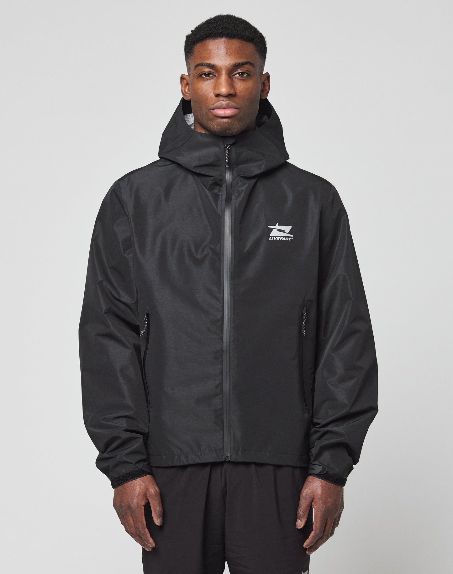 A man stands facing forward against a plain light background, wearing the LIVE FAST DIE YOUNG Live Fast Performance Rain Jacket in black, with a regular fit, zip-up front, and white logo on the chest. His hands rest relaxed by his sides.