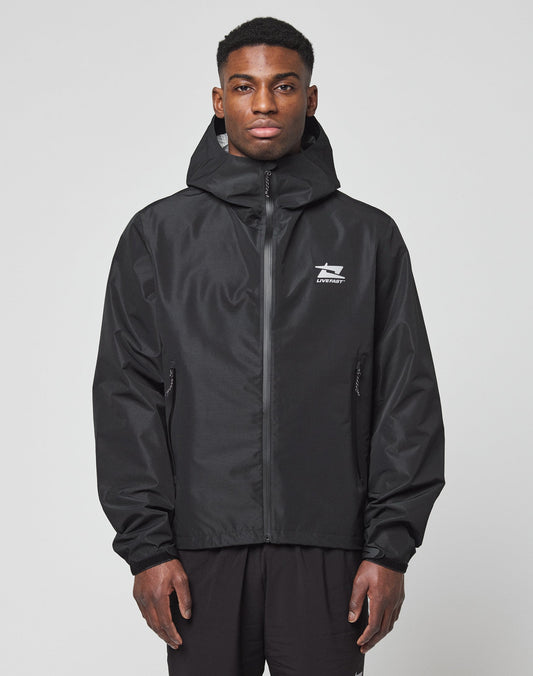 A man stands facing forward against a plain light background, wearing the LIVE FAST DIE YOUNG Live Fast Performance Rain Jacket in black, with a regular fit, zip-up front, and white logo on the chest. His hands rest relaxed by his sides. | model