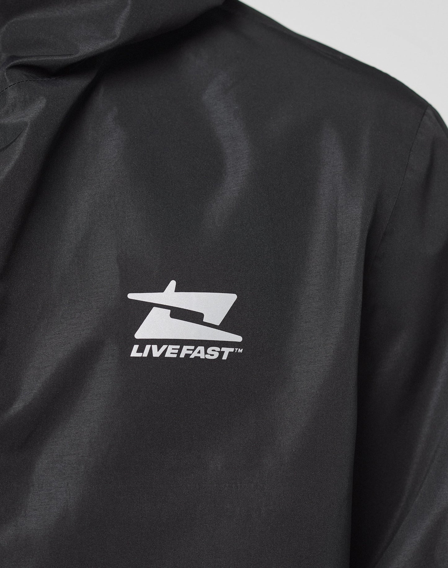 Close-up of the LIVE FAST DIE YOUNG Live Fast Performance Rain Jacket in black, featuring a white graphic logo and LIVE FAST™ on the chest. The image highlights the logo, shoulder area, and its Regular Fit for comfort.