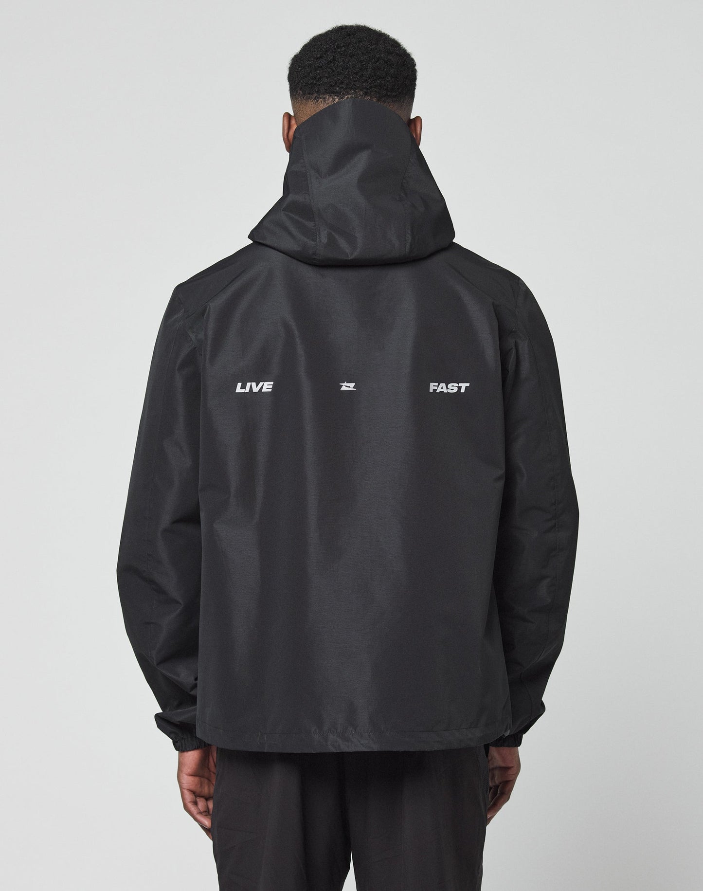 Seen from behind, a person wears the LIVE FAST DIE YOUNG Live Fast Performance Rain Jacket in black, featuring wasserdichte Nähte, a Kapuze mit Visor, and LIVE and FAST in white on the back against a plain white background.