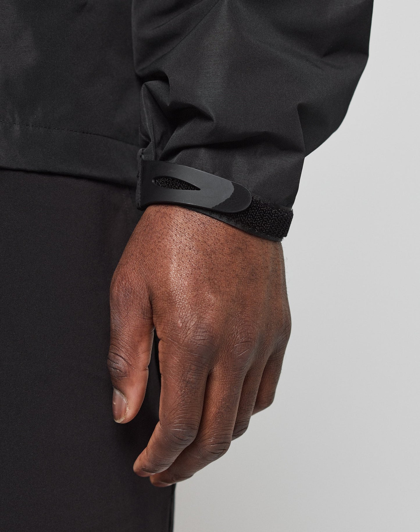 A close-up shows a persons hand and wrist wearing the LIVE FAST DIE YOUNG Live Fast Performance Rain Jacket with waterproof seams and an adjustable Velcro sleeve cuff, set against a plain, light-colored background.