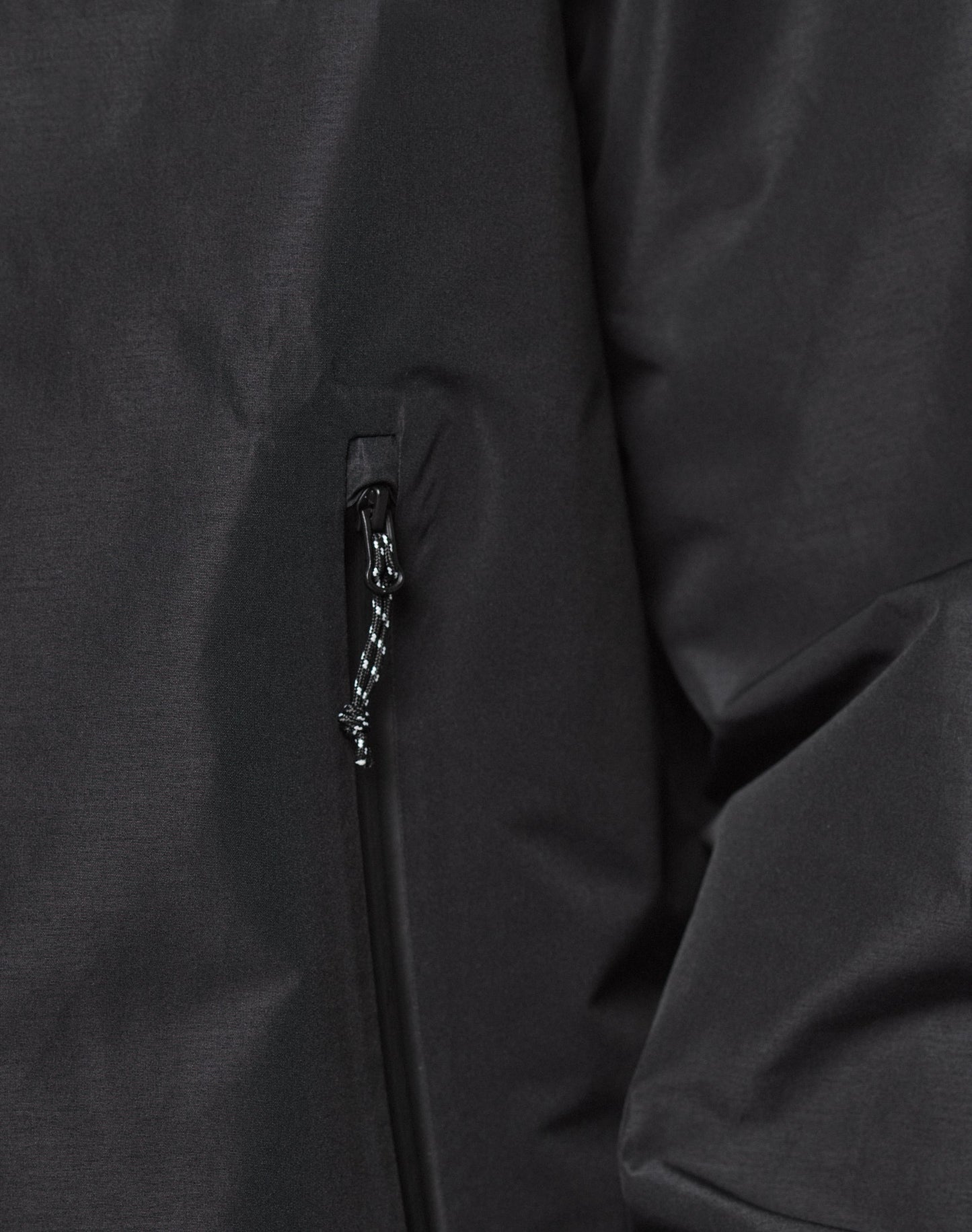 Close-up of LIVE FAST DIE YOUNG’s Live Fast Performance Rain Jacket in black, highlighting a zippered pocket with black and white pull cord, partial view of the sleeve and body fabric with elastic cuff.