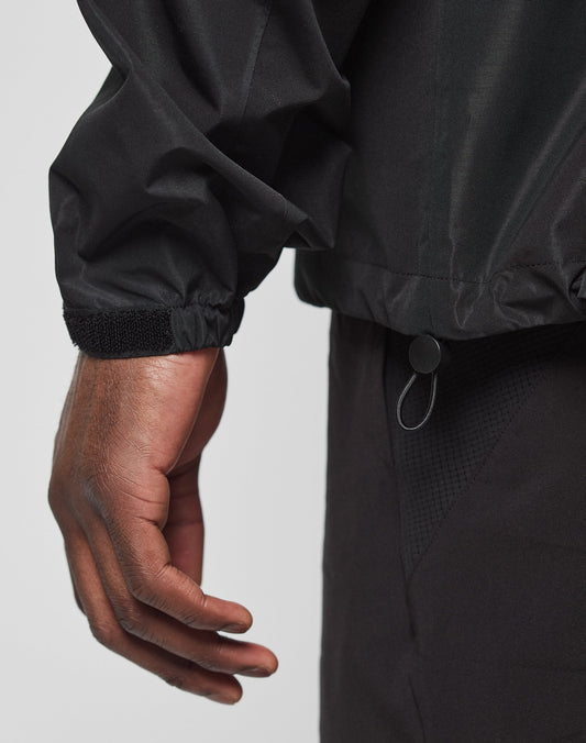 A close-up shows a person’s left hand and arm wearing the LIVE FAST DIE YOUNG Live Fast Performance Rain Jacket with elastic cuffs, an adjustable hem drawstring, regular fit, and black pants against a plain light background.