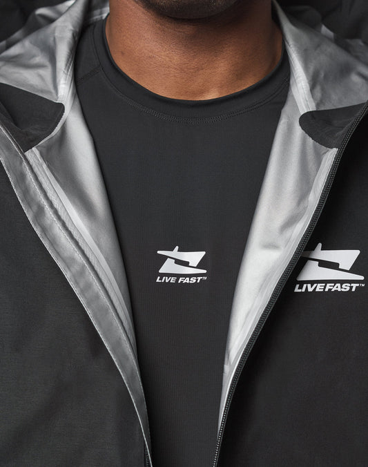 Close-up of a person wearing the black Live Fast Performance Rain Jacket by LIVE FAST DIE YOUNG, featuring Elastische Ärmelöffnung and partially unzipped to reveal a black shirt with the LIVE FAST logo and arrow design.