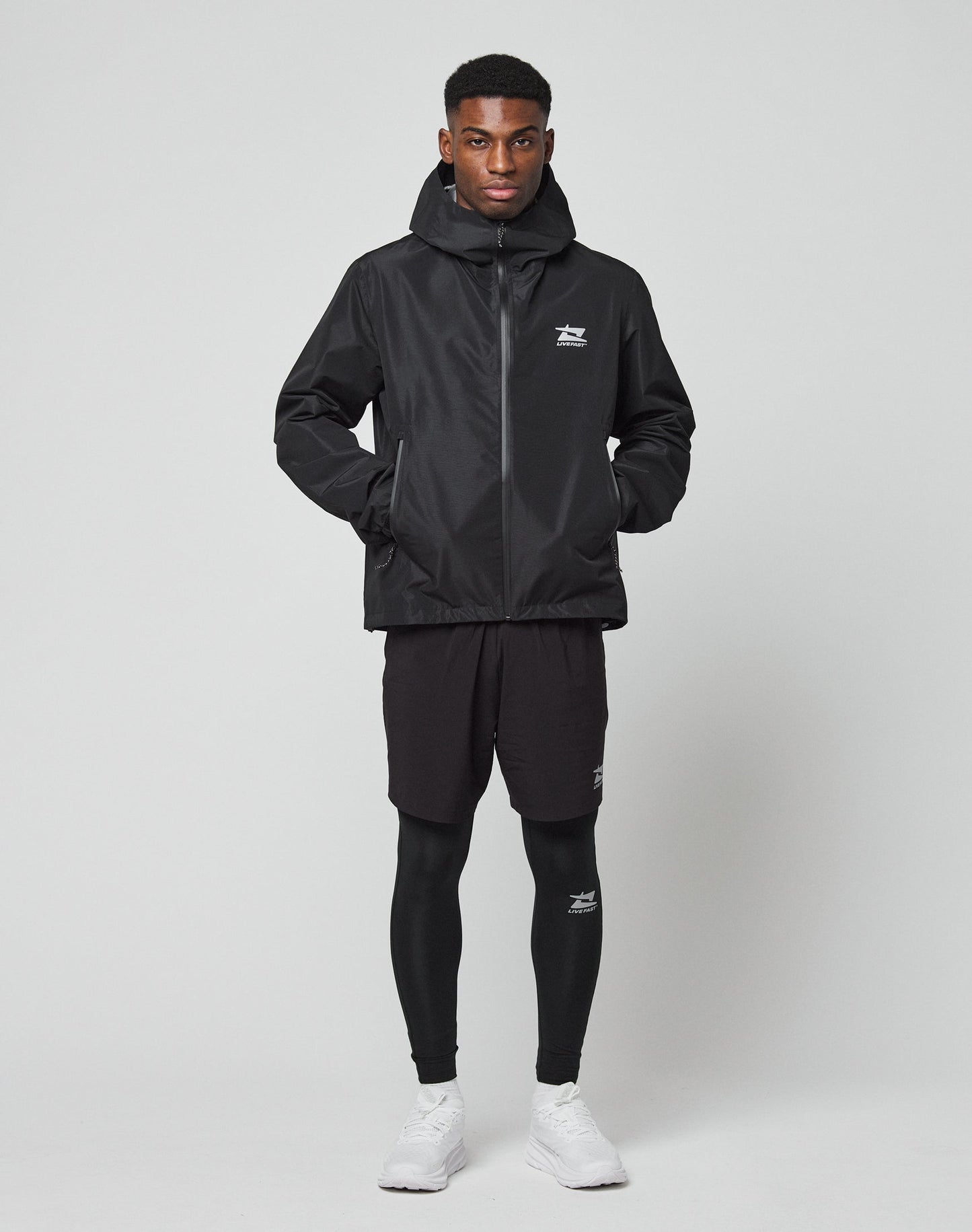 A man stands facing forward wearing LIVE FAST DIE YOUNG Live Fast Performance Tights under black shorts, a slim fit black hooded jacket, and white athletic shoes with subtle Live Fast branding, set against a plain light gray background.