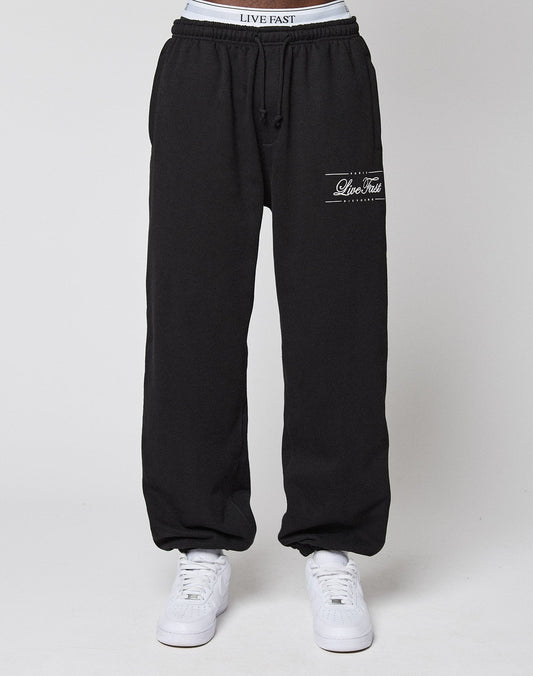 A person wearing black Wide Cuff Jogger sweatpants by LIVE FAST DIE YOUNG, featuring a Live Fast logo on the left thigh, a comfortable elastic waistband, soft French Terry fabric, and paired with white sneakers against a plain white background.