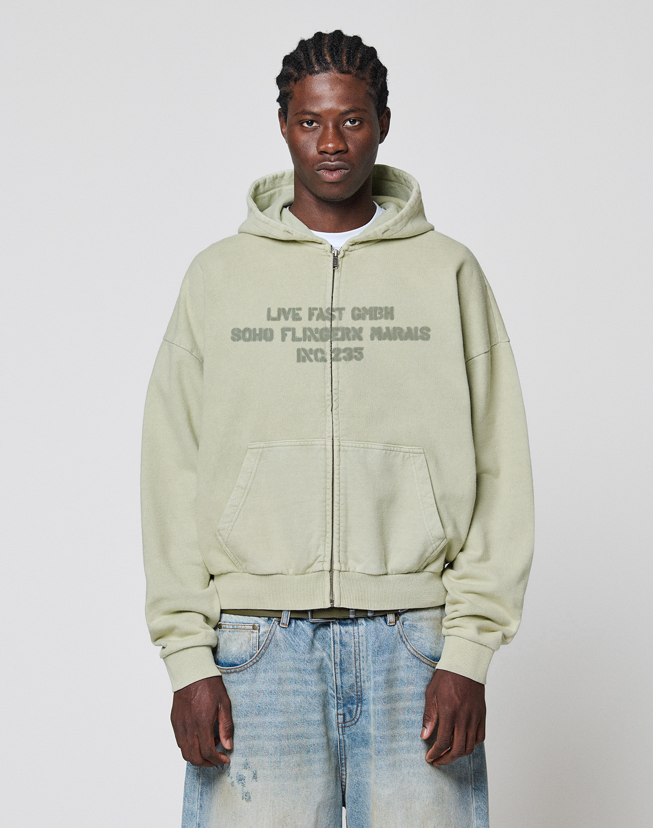 A man with braided hair wears the LIVE FAST DIE YOUNG LFDY District Hooded Zip in light green, featuring a front print, paired with loose-fitting, faded oversized jeans, standing against a plain white background. The hoodie is 100% Baumwolle.