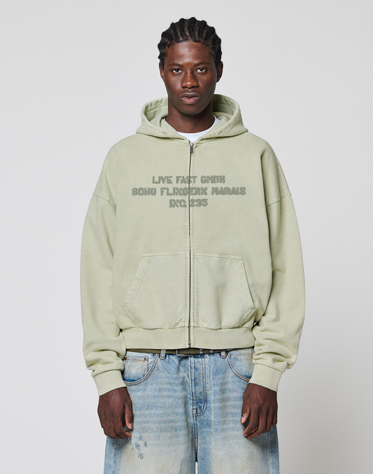 A man with braided hair wears the LIVE FAST DIE YOUNG LFDY District Hooded Zip in light green, featuring a front print, paired with loose-fitting, faded oversized jeans, standing against a plain white background. The hoodie is 100% Baumwolle. | model