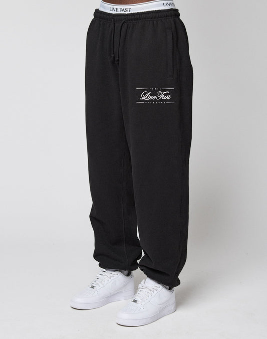 A person wears black Wide Cuff Joggers by LIVE FAST DIE YOUNG, featuring "Live Fast" on the left thigh, elastic waistband, and cuff. Made from soft French Terry. Styled with white sneakers against a plain light background. | model