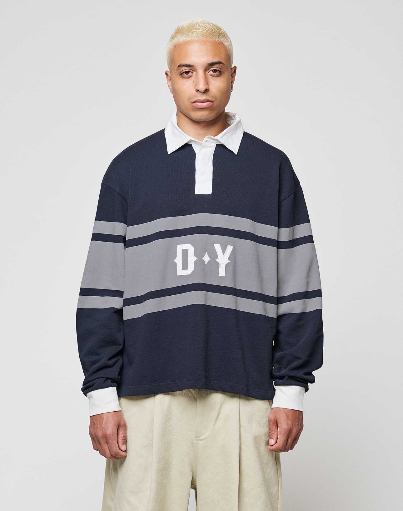A person with short bleached blond hair wears a navy and gray LFDY Striped Polo by LIVE FAST DIE YOUNG, featuring "D*Y" on the front, paired with beige cotton twill pants, standing against a plain light gray background. | model