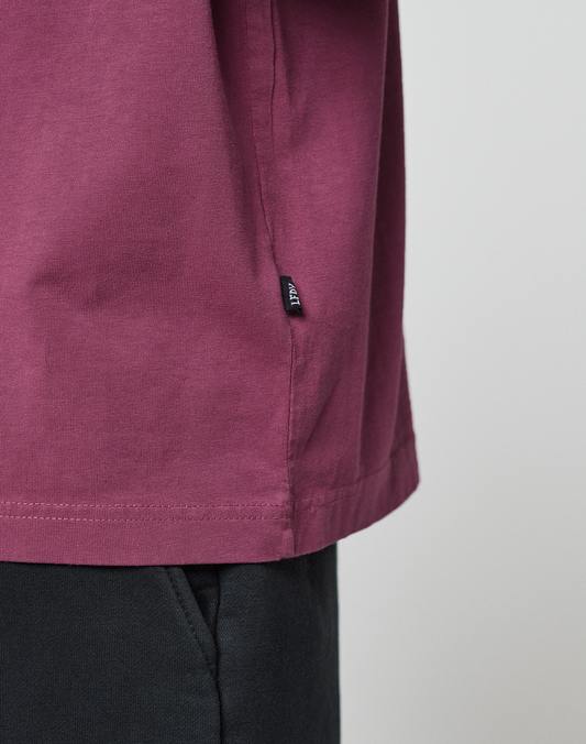 Close-up of a person wearing the LIVE FAST DIE YOUNG Basic 210 Tee in maroon, featuring an oversized fit and a small black tag on the side seam, paired with black pants. The background is plain and light-colored.