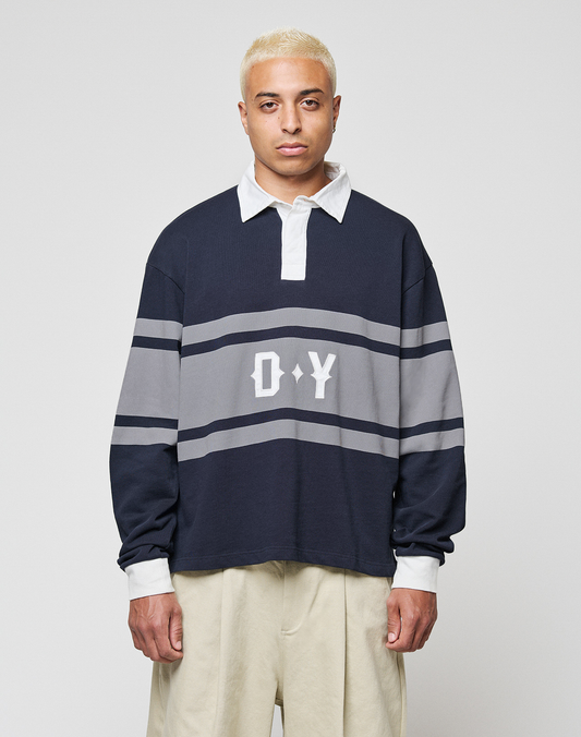 A person with short bleached blond hair wears a navy and gray LFDY Striped Polo by LIVE FAST DIE YOUNG, featuring "D*Y" on the front, paired with beige cotton twill pants, standing against a plain light gray background. | model