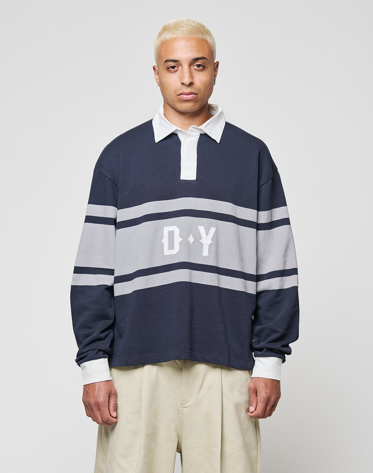 A person with short bleached blonde hair wears the LIVE FAST DIE YOUNG LFDY Striped Polo, an oversized navy and gray long-sleeve shirt with white collar and cuffs, D Y on the chest, and loose beige cotton-twill pants, against a plain background.