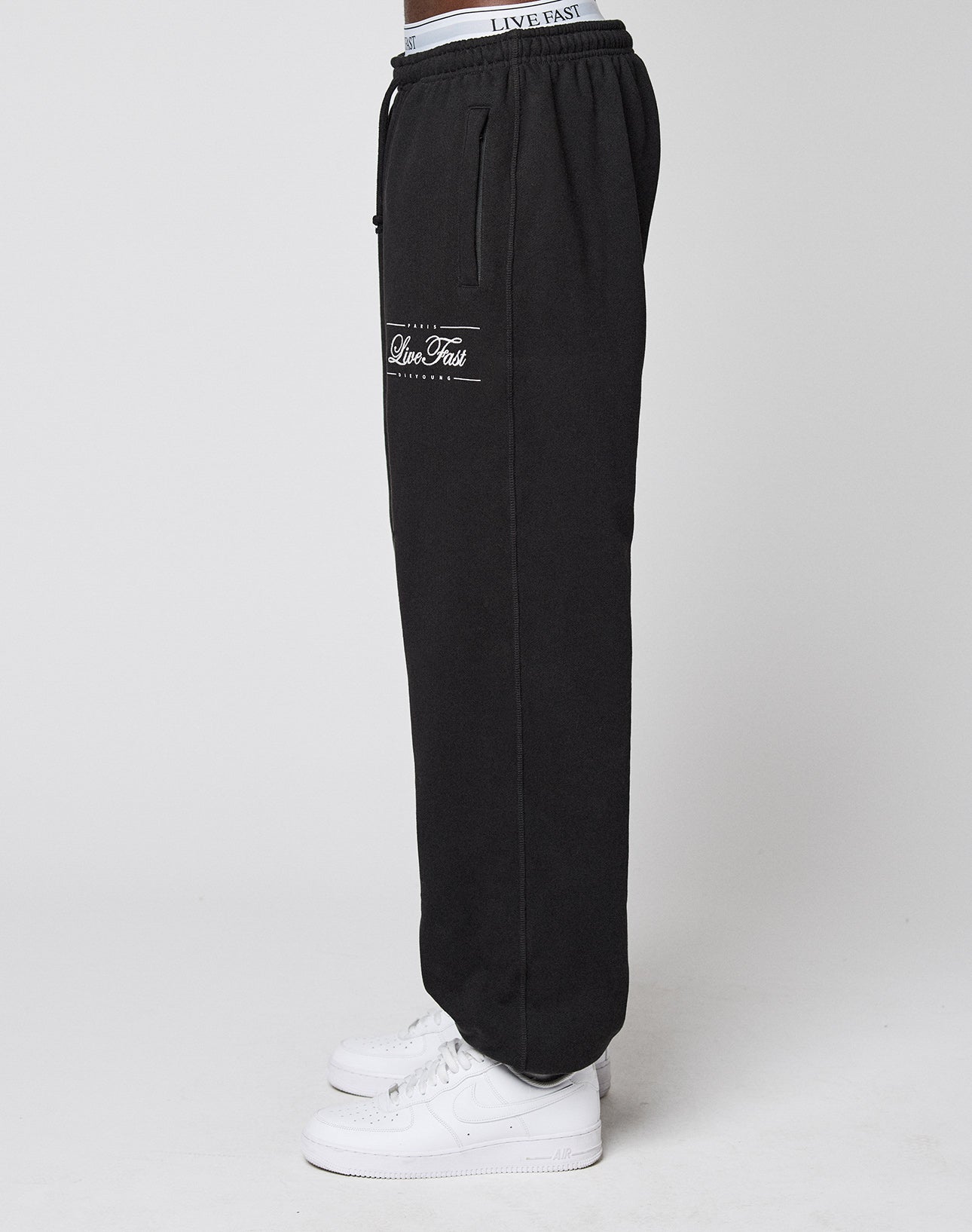 A person stands sideways in LIVE FAST DIE YOUNG Wide Cuff Joggers with “Live Fast” on the pocket, white sneakers, and a visible LIVE FAST waistband, set against a plain white background.