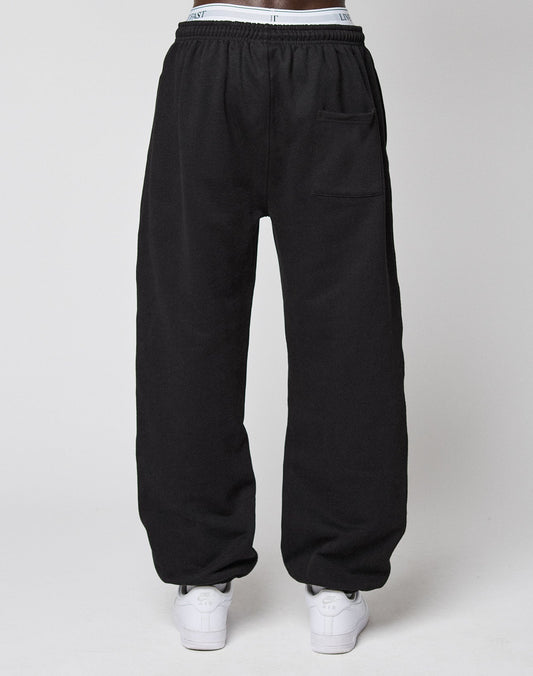 A person wearing LIVE FAST DIE YOUNG Wide Cuff Jogger in black, featuring an elastic waistband and a right thigh pocket, paired with white sneakers, stands against a plain white background.