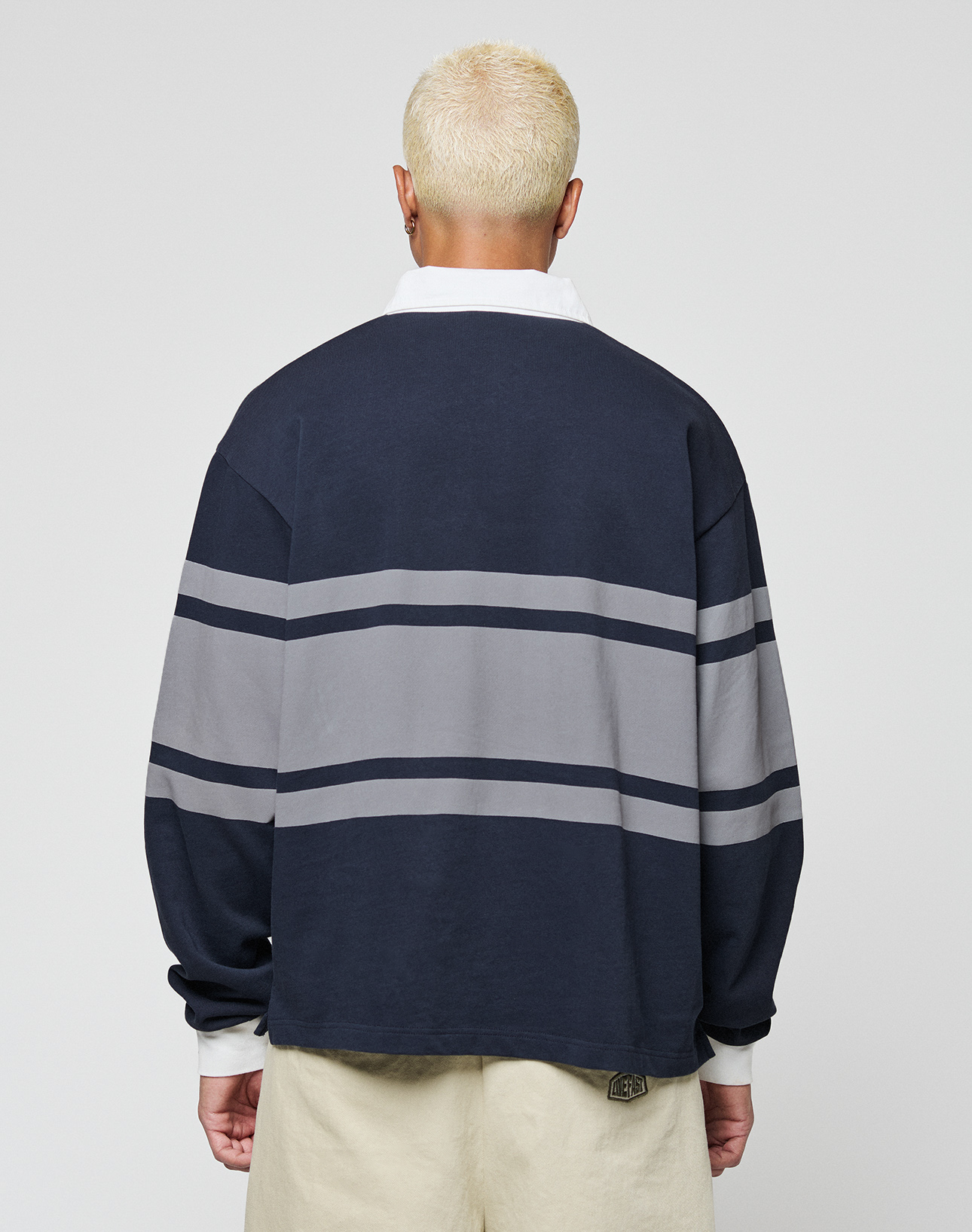 A person with short, bleached blonde hair is seen from behind in a LIVE FAST DIE YOUNG LFDY Striped Polo—an oversize fit navy sweater with two gray stripes, white collar and cuffs—paired with beige cotton-twill pants, against a plain light background.