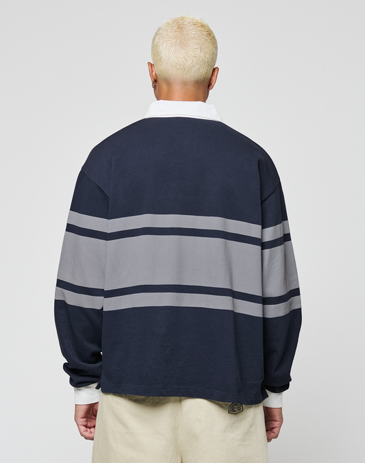 A person with short, bleached blonde hair is seen from behind in a LIVE FAST DIE YOUNG LFDY Striped Polo—an oversize fit navy sweater with two gray stripes, white collar and cuffs—paired with beige cotton-twill pants, against a plain light background.