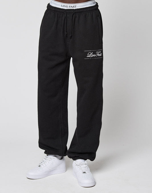 A person wears black Wide Cuff Joggers by LIVE FAST DIE YOUNG with a visible French Terry waistband and "LIVE FAST" text, paired with white sneakers, standing against a plain white background.