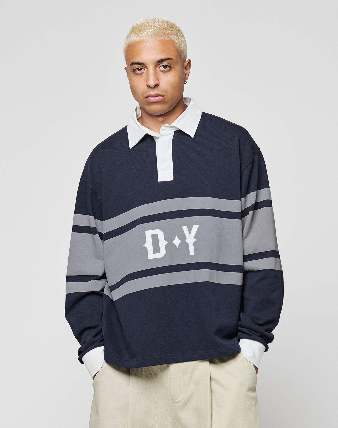 A person with short bleached blonde hair wears the LFDY Striped Polo by LIVE FAST DIE YOUNG, layered over a white collared shirt with beige pants, standing against a plain light background.
