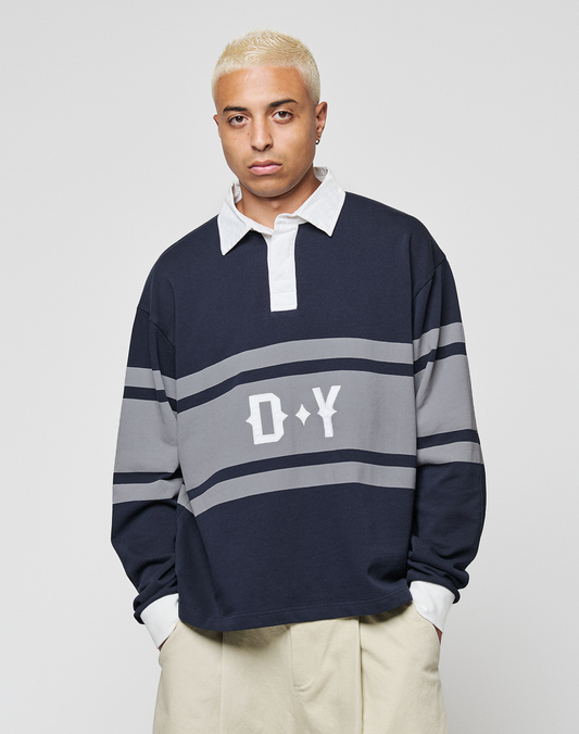 A person with short bleached blonde hair wears the LFDY Striped Polo by LIVE FAST DIE YOUNG, layered over a white collared shirt with beige pants, standing against a plain light background.